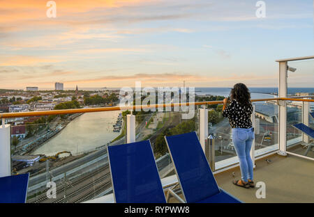 Eine Frau auf dem oberen Deck eines Kreuzfahrtschiffes genießt den Sonnenuntergang über der Stadt am Meer, Strand, Ostsee, Alten Strom Kanal und Bahn in Warnemünde Deutschland Stockfoto