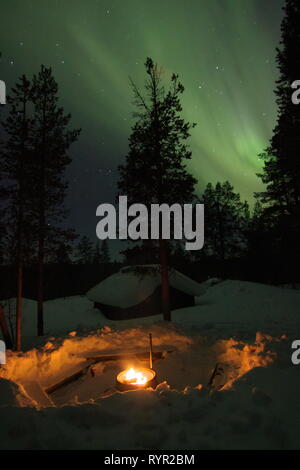 Kamin mit einem Holzhaus und Bäume im Schnee hinter und Aurora Borealis (Nordlicht) oben in einer Winternacht im finnischen Lappland. Saar Stockfoto