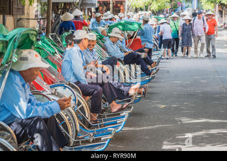 Hoi An, Vietnam - 23. Oktober 2018: Eine lange Reihe von trishaws in Uniformen warten auf Kunden, die Annäherung der Gruppe der Touristen in konischen Hüten. Stockfoto