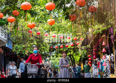 Hoi An, Vietnam - Oktober 23, 2018: eine Straße mit Perlen von Laternen hängen von Vordach von üppigem Laub und eine reife Frau in Kleid & ein Mädchen auf dem Fahrrad. Stockfoto