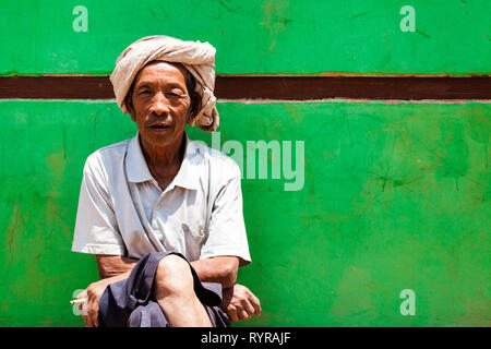 Eine traditionelle burmesische Mann sitzt vor einer grünen Wand in Kalaw, Myanmar Stockfoto