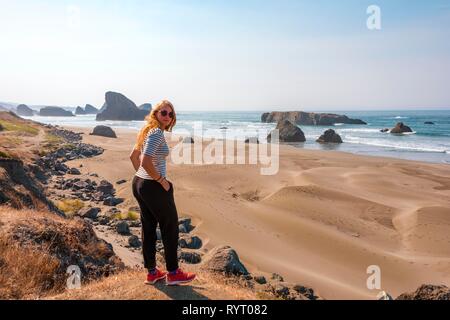 Junge Frau in die Kamera schaut, Sandstrand und an der Küste mit schroffen Felsen, Myers Creek Strand Sicht, Oregon USA Stockfoto