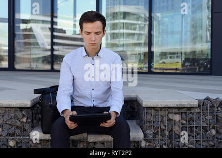 Junge Kerl in Freelancer shirt arbeitet auf der Straße in der Nähe der Gebäude der Stadt. Der Mann hält Tablet. nächsten Tasche Aktentasche Stockfoto