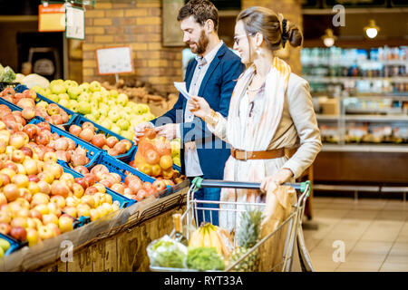 Junge und glückliche Paar kaufen frische Früchte zusammen mit Einkaufsliste und Warenkorb im Supermarkt Stockfoto