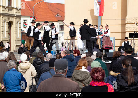 Sopron, Ungarn. 15 Mär, 2019. Gruppe männlicher Volkstänzer führt Traditionelle Ungarische Tänze im Kreis auf der Bühne Petőfi Square, Sopron, Ungarn. Mädchen der Volkstanzgruppe zusammen Klammern am Rand der Bühne beobachten. Credit: Wahavi/Alamy leben Nachrichten Stockfoto