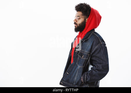 Profil von verträumt und nachdenklich schöne stilvolle african american Kerl in Denim Jacke über rot Hoodie, halten sich an den Händen in den Taschen als Stockfoto