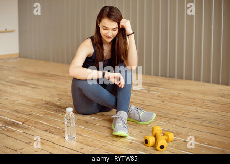 Junges Mädchen mit dunklem Haar sitzt auf dem Boden und richtet ihre Fitness tracker Training im Fitnessraum Stockfoto