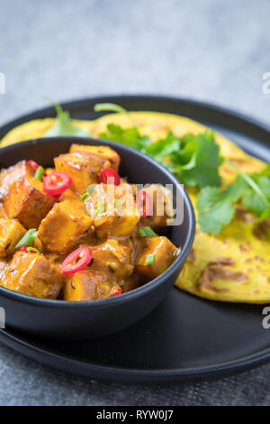 Curry und Roti close up auf einer schwarzen Platte und einen grauen Hintergrund. Masala paneer Curry in eine Schüssel mit frischem Roti und Koriander auf schwarzem Teller. Bunte Stockfoto