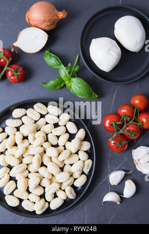 Frischen Mozzarella mit Ungekochte Gnocchi auf einem schwarzen Platte und einem dunkelgrauen Hintergrund. Italienischen Zutaten für gebackene Gnocchi. Reben fröhlichen Tomaten, f Stockfoto