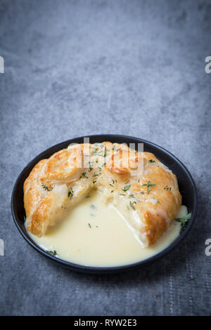 Nässen Camembert im Teigmantel. Geschmolzener Camembert mit Verkrusteten puff Pasty und Kräuter auf einer schwarzen Platte. Grauer Hintergrund. Traditionelle französische Käse. Stockfoto