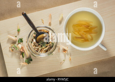 Tasse Zitronengras Tee und ein Glas Glas mit getrockneten Blätter von zitronengras. Holz und Papier Hintergrund. Ansicht von oben. Stockfoto