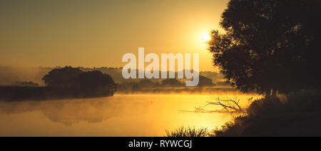 Die Konturen der Häuser des Dorfes ertrinken in dem dichten Nebel der Dämmerung am Morgen über die hellen und orange Landschaft der Stockfoto