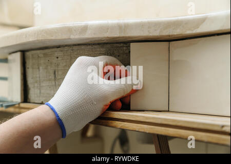 Die Hand des Arbeitnehmers setzt das Stück der Fliese auf der gekrümmten Wand unter einem Waschbecken. Stockfoto