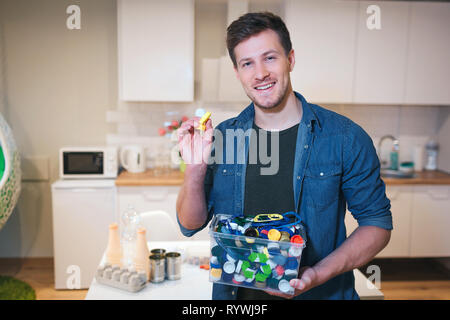Die Umwelt zu schützen. Kunststoff Deckel bereit für das Recycling. Junge lächelnde Mann, Recycling Container mit Deckel Flaschen auf Küche gefüllt Stockfoto