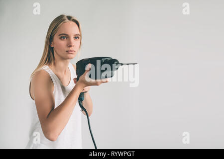 Lustige portrait umgekippt Handwerkerin. Blonde Frau trägt Shirt isoliert auf Weiss. Mädchen, das nicht weiß, wie man eine Bohrmaschine zu benutzen Stockfoto