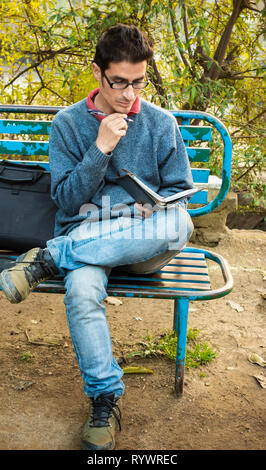 Outdoor Bild von asiatischen, indischen serene junger Erwachsener Geschäftsmann lesen Business Planner und auf einer Parkbank am Tag Zeit sitzen. Stockfoto