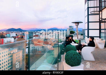 Ljubljana, Slowenien - 14. Januar 2019: Paar an Street Cafe Terrasse mit Tischen und Stühlen und herrlichem Panoramablick in Ljubljana Altstadt, Sonnenuntergang, Slowenien. Menschen im modernen Restaurant Veranda Stockfoto