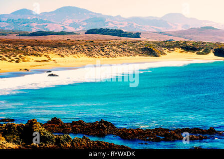 Mit Blick auf die Felsen, Klippen und goldene Sandstrände, blaues Meer mit Wellen auf Ufer mit Dunstige Berge unter hellen dunstigen Himmel brechen. Stockfoto