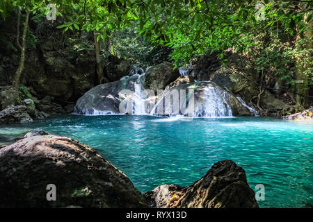 Grüne Blätter an der Spitze und dunklen Felsen im vorderen Rahmen eine schöne kurze Wasserfall mit glatten Felsen im Wald der Erawan Nationalpark in Thailan Stockfoto