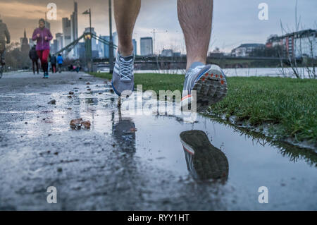 Blick auf die Beine eines Mannes, die nach dem Regen entlang eines Flusses in einer großen Stadt. Frühling Sport Stockfoto