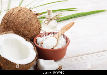 Hintergrund der Kokosnuss, Kokosnussschalen, Öl in Ton Schüssel und Glas Glas und Löffel auf weiße Holztisch Stockfoto