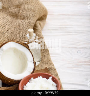 Kokosöl in braun Ton Schale, Glas, Flasche, Kokosnussschalen mit Fleisch auf Hanf Sack auf weißen Küche aus Holz Tisch Stockfoto