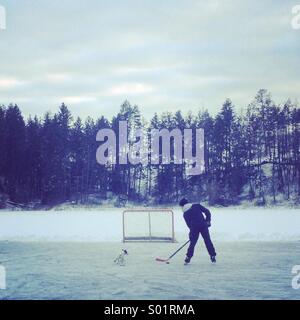 Kanadische Landschaft: ein Junge spielt Hockey auf einem zugefrorenen Teich mit seinem Hund im Schlepptau. Stockfoto