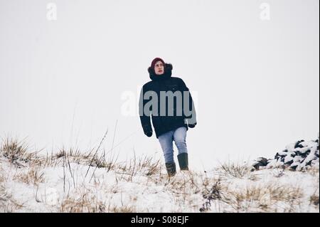 Ein Teenager arbeitet durch ein schneebedecktes Feld im Nordosten von England in einem schwarzen Parka Mantel und Gummistiefel. Stockfoto