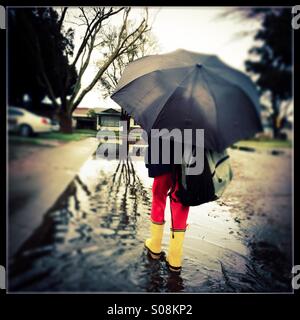 Ein sechs Jahre alter Junge mit einer Schultasche durchschreitet hält einen Regenschirm und Gummistiefel tragen einer überfluteten Straße auf dem Weg zur Schule. Stockfoto