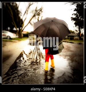 Ein sechs Jahre alter Junge mit einer Schultasche durchschreitet hält einen Regenschirm und Gummistiefel tragen einer überfluteten Straße auf dem Weg zur Schule. Stockfoto