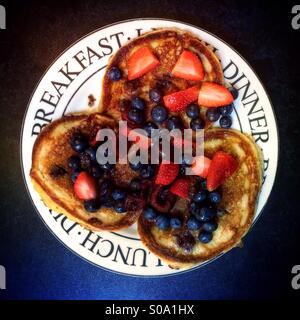 Amerikanische Pfannkuchen mit Ahornsirup, Erdbeeren, Kirschen und Blaubeeren auf einem weißen Teller gegen eine schwarze Fläche. Stockfoto