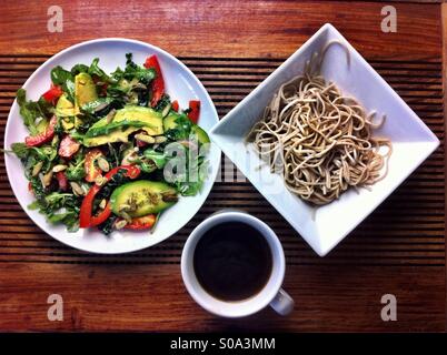 Veganes Mittagessen - Soba-Nudeln in quadratische Schüssel, Salat auf Runde weiße Platte und Kaffee in weiße Tasse auf einem dunklen Holz Tablett serviert Stockfoto