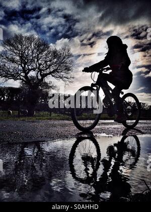 Junges Mädchen auf dem Fahrrad in der Silhouette spiegelt sich in der Pfütze Stockfoto