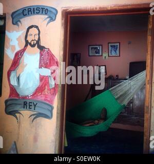 Ein älterer Mann schläft auf einer Hängematte in seinem Haus verziert mit einem Gemälde von Jesus Christus in Santa Elena, Yucatan, Mexiko Stockfoto