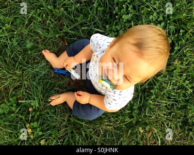 Blick von der Spitze eines 8 Monate alten lächelnden Baby jungen draußen sitzen auf dem Boden, grünen Rasen mit Schnuller im Mund, hält eine Wäscheklammer Stockfoto