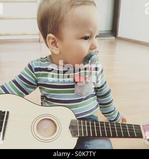 Baby Junge Gitarre spielen, Blick auf die Seite, sitzend auf einem Holzfußboden in einem leeren Raum mit einem Schnuller im Mund Stockfoto