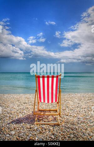 Ein einzelnes rot und weiß gestreiften Liegestuhl auf einem Kiesstrand direkt am Meer an einem schönen Sommertag. Stockfoto