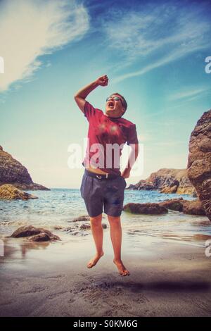 Ein Junge auf einem sandigen Strand springen vor Freude. Ein Cornish Strand mit dem Meer hinter ihm. Stockfoto