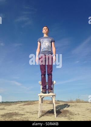 Mann in Hemd und Hose auf einem Stuhl auf dem Hügel, die ernsthaft auf der Suche in die Ferne auf blauen Himmelshintergrund Stockfoto