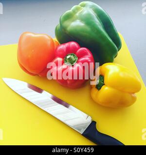 Eine rote, orangefarbene, gelbe und grüne Paprika mit einem Messer auf einen gelben Hackstock. Stockfoto