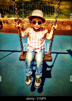 Ein kleiner Junge mit Hut und Sonnenbrille spielt auf s Schaukel im Park. Stockfoto