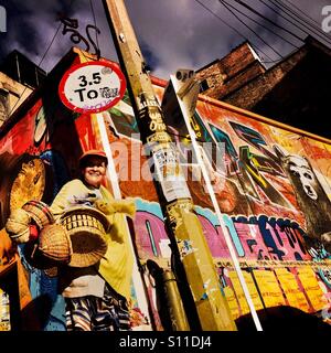 Eine kolumbianische Straße Verkäufer Spaziergänge rund um eine Straßenecke, bedeckt mit bunten Graffiti-Kunstwerk und Plakate in La Candelaria, Bogota, Kolumbien, 19. Februar 2016. Stockfoto