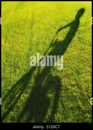 Der lange Schatten eines 6 Jahre alten Jungen auf seinem Fahrrad in eine offene Grasfläche. Ein Bild mit Bereich für Kopie. Bildnachweis - © COLIN HOSKINS. Stockfoto