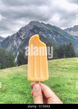 Eis am Stiel in einer männlichen kaukasischen Hand vor einem Berg und einer grünen Wiese in den österreichischen Alpen Stockfoto