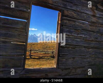 Blick auf die Teton und den Grand Teton National Park und Zaun durch das Fenster eine alte verlassene Blockhütte. Stockfoto