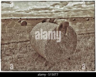 Ein sieben Jahre alter Junge liegt auf einer frisch hergestellten Heuballen in ein Feld, das vor kurzem geerntet worden ist. Schläft der junge? Spielt er? Wo sind seine Freunde? Eltern? Bildnachweis - © COLIN HOSKINS. Stockfoto