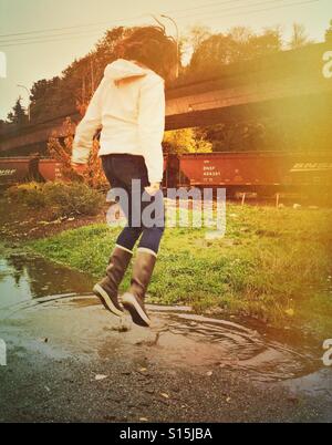 Eine verspielte Frau trägt Regen Stiefel Sprünge in einer Pfütze Regen an einem Herbsttag im pazifischen Nordwesten. Stockfoto