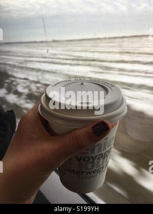 Hand hält eine Tasse Kaffee in einem fahrenden Zug und mit Blick auf die Landschaft aus dem Fenster hinter gehen Stockfoto