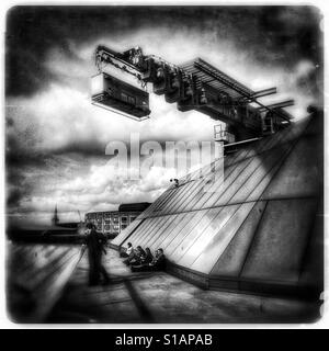Schwarz / weiß Foto mit einem futuristischen, traumhafte Gefühl von Büroangestellten sitzen, wie ein große Service-Kran über Kopf bewegt sich auf der Dachterrasse eine neue Änderung, London, England, UK. Stockfoto