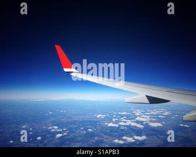 Blick durch die Fenster ein Flugzeug von einem Verkehrsflugzeug Flügel mit einer hellen roten Flügelspitze, fliegen hoch über den Wolken vor einem tiefblauen Himmel. Stockfoto